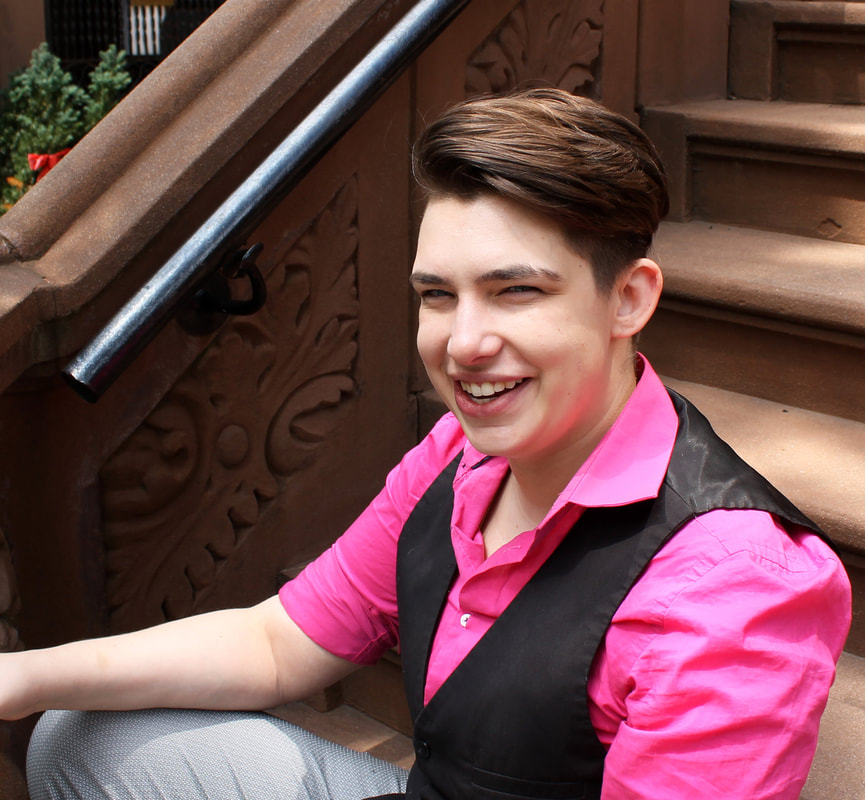headshot of Aiden Feltkamp, a smiling white trans nonbinary person in a pink shirt with a black vest sitting on steps in new york city